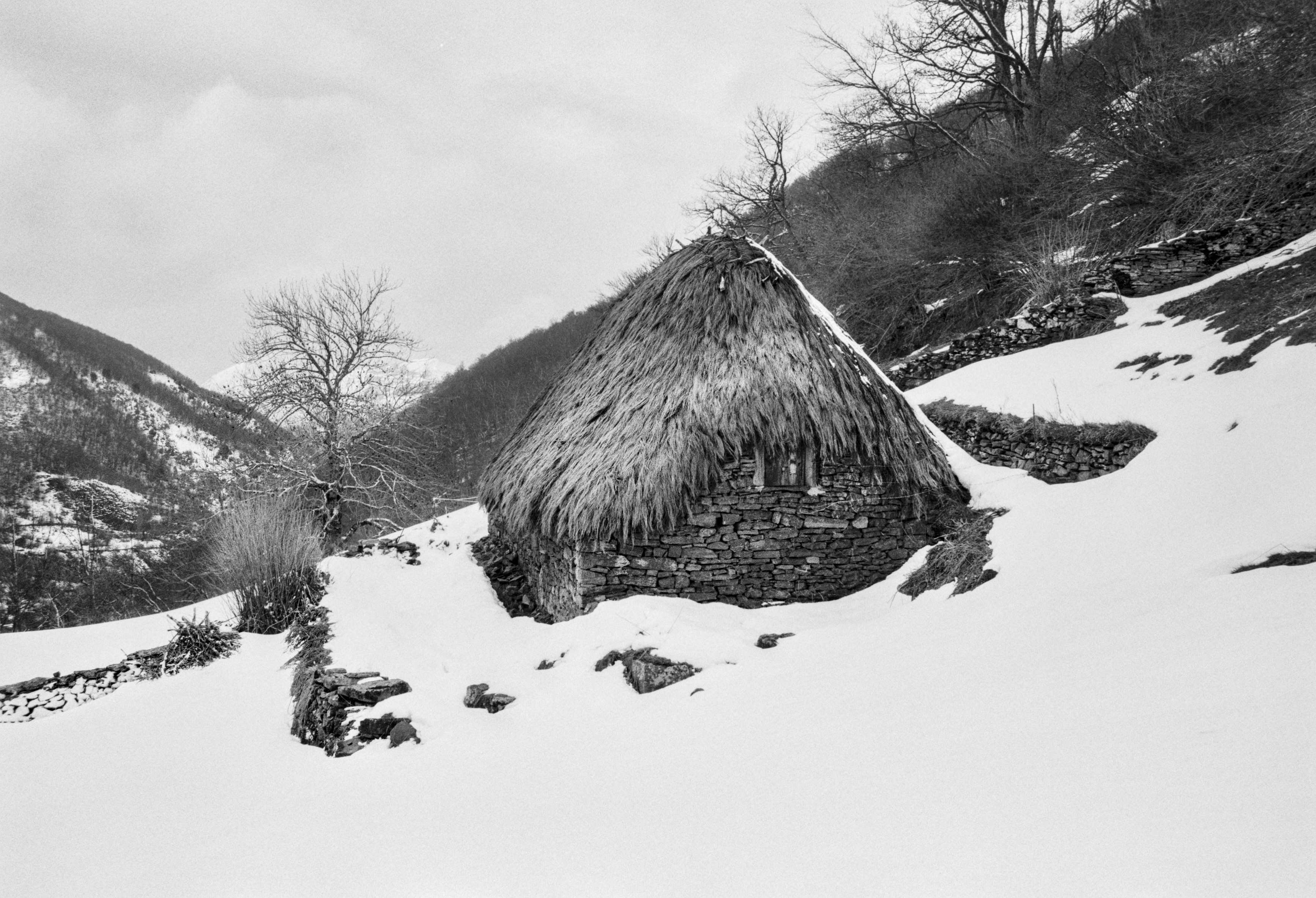 A black and white photograph of a traditional stone palloza with a thatched roof surrounded by snow, set against snowy mountains and bare winter trees.
