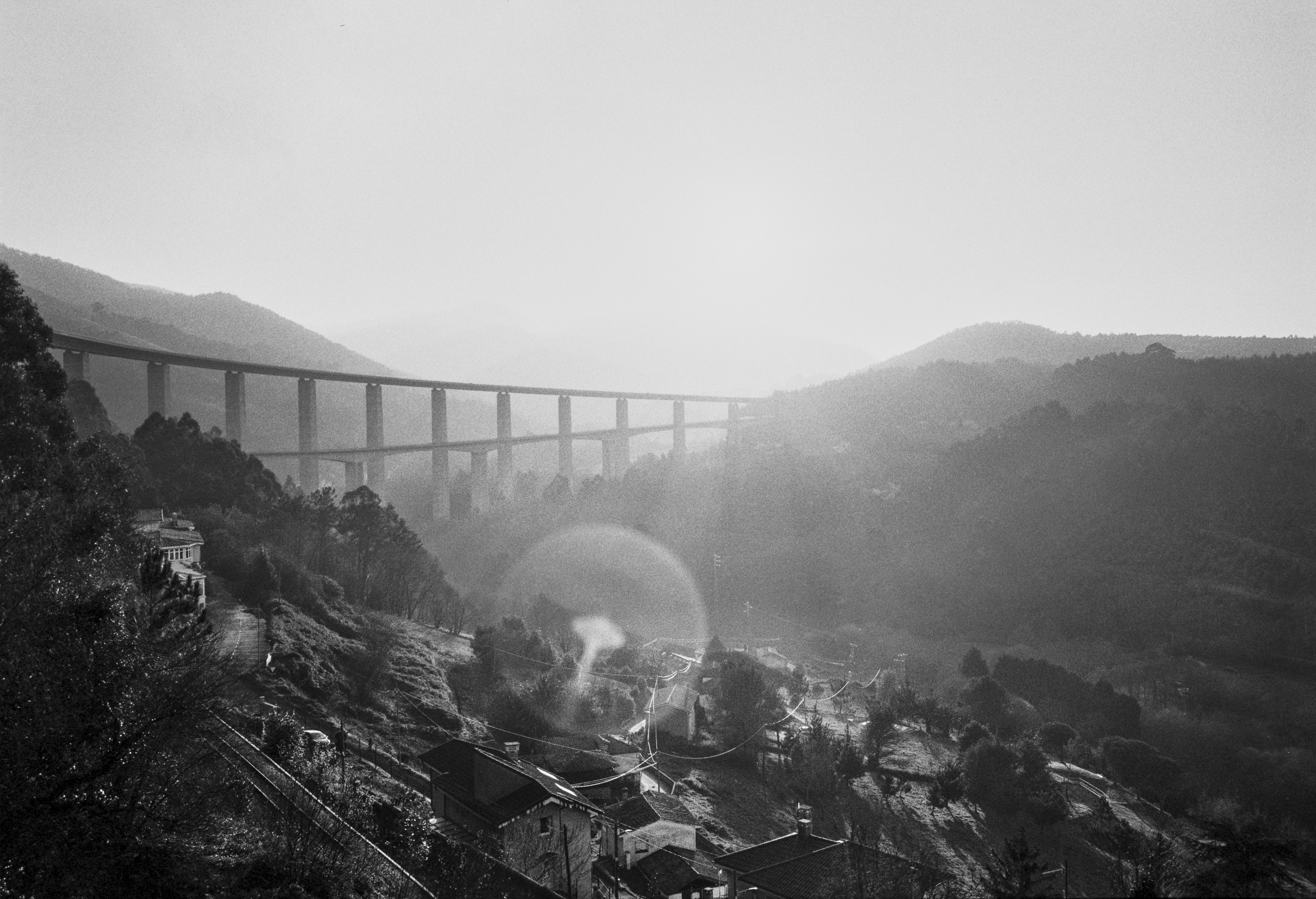 A black and white photograph of a valley at sunset with a tall viaduct crossing the scene, houses on the hillside below and hazy light filling the landscape.