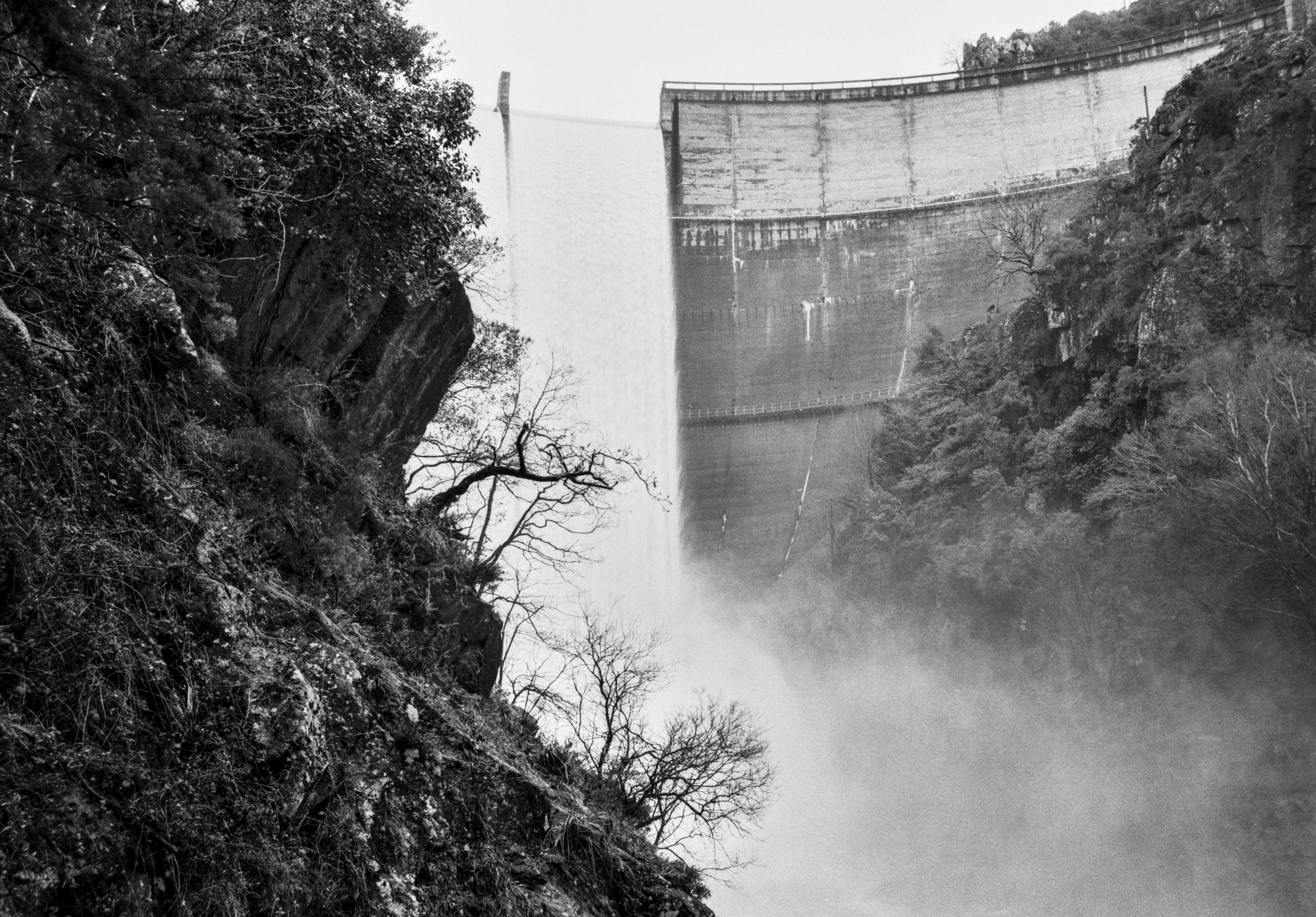 A black and white photograph of a dam releasing water in a powerful vertical stream, with rocky cliffs and trees framing the scene and mist rising below.