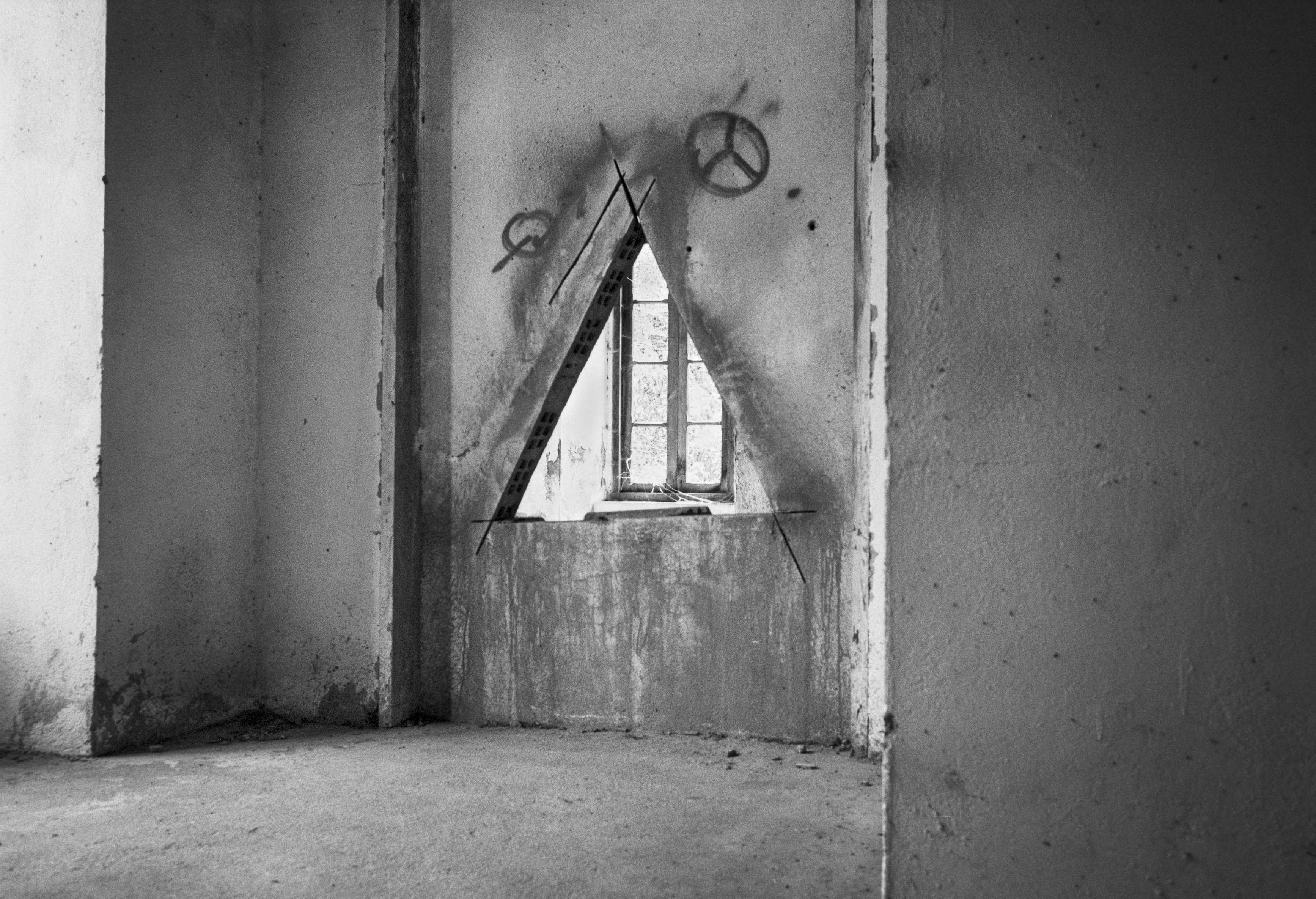 A black and white photograph of an unfinished or abandoned interior room with a triangular opening cut into a wall, revealing a window behind it. Peace symbol graffiti is visible above the opening.
