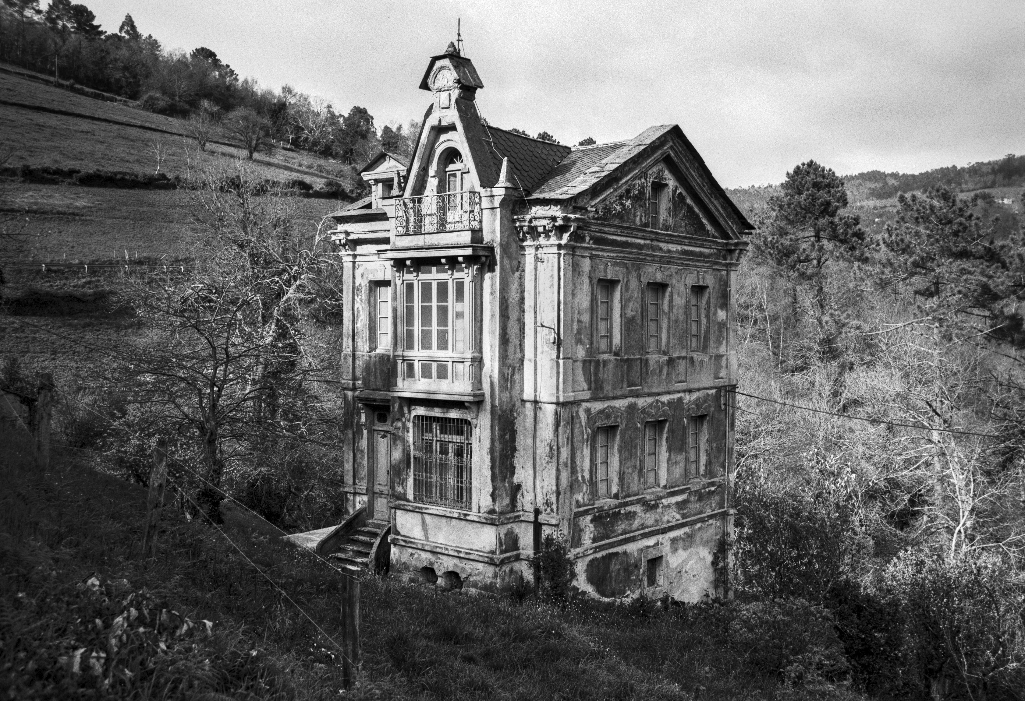 A black and white photograph of an old three story indiano style house with weathered walls, surrounded by fields, trees and a hillside.