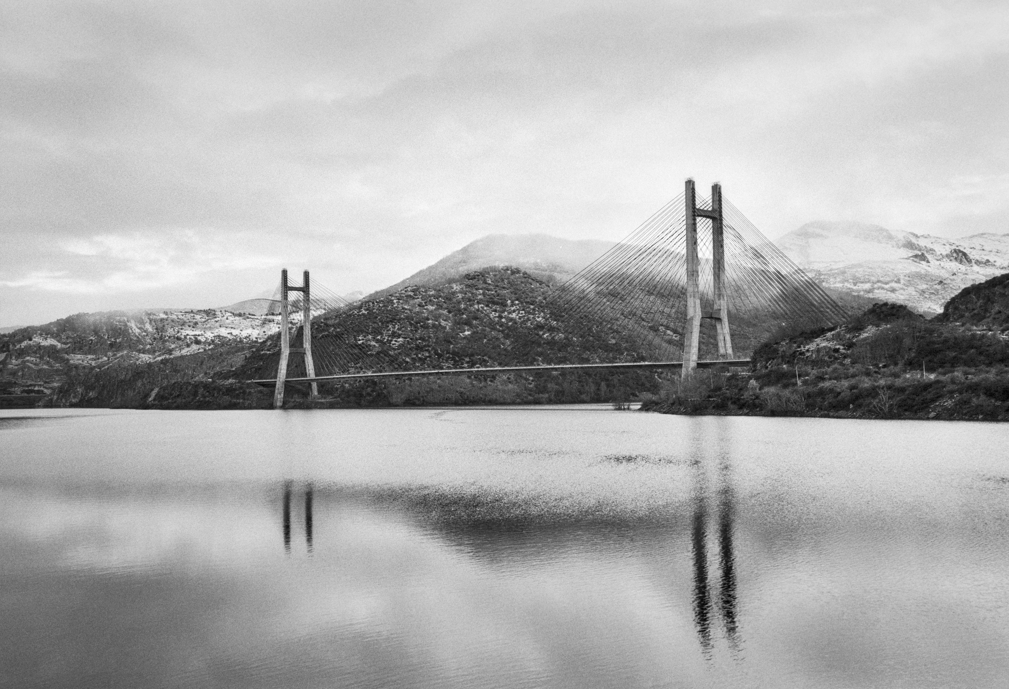 A black and white photograph of a large cable stayed bridge crossing a calm reservoir, with mountains in the background and reflections in the water. The scene is viewed from a distance on a cloudy day.