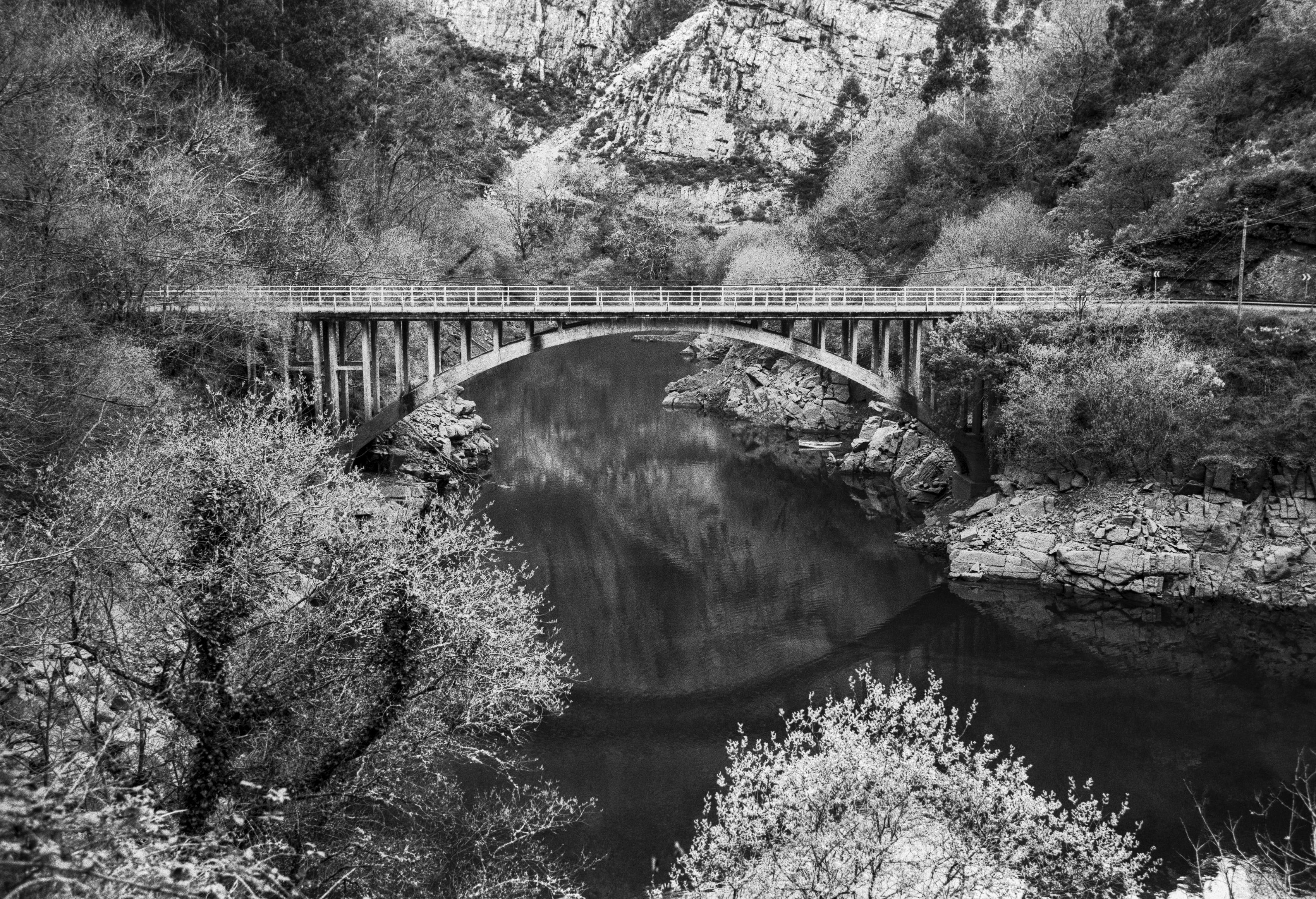 A quiet arched bridge crossing a dark, reflective river surrounded by steep mountains and early spring foliage.