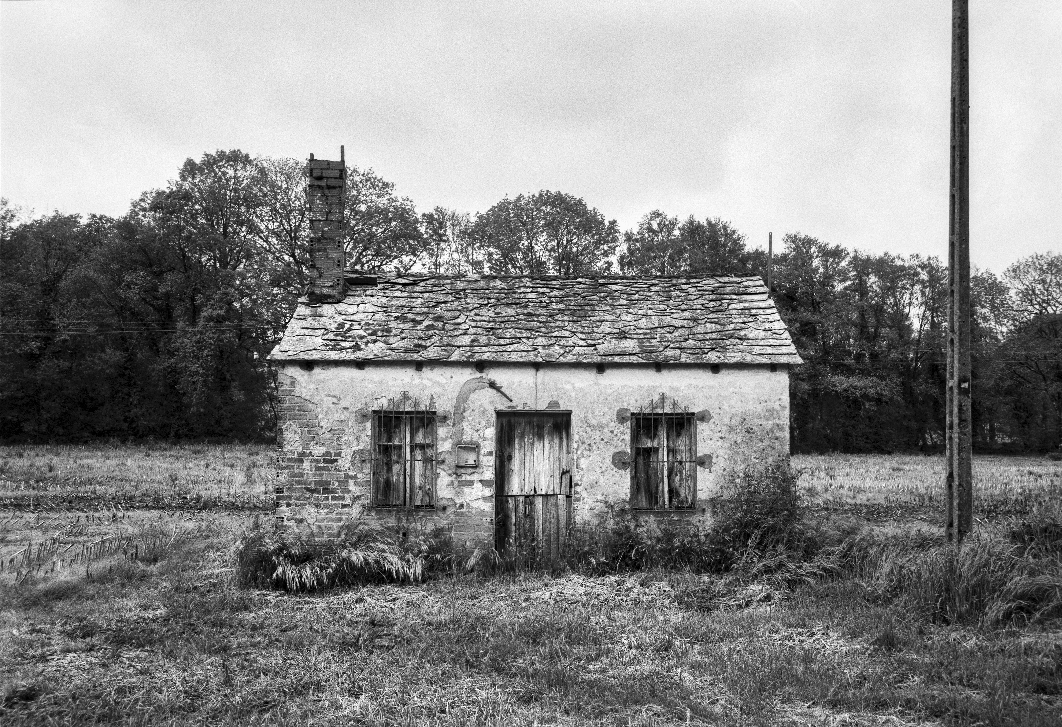 A small, weathered stone house stands alone in a field, with barred windows and a cracked wooden door.
